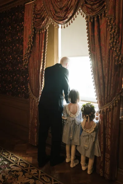 A tender flower girls moment, a candid wedding moment with two girls in matching dresses and a floral crown, at a window with damask wallpaper.
