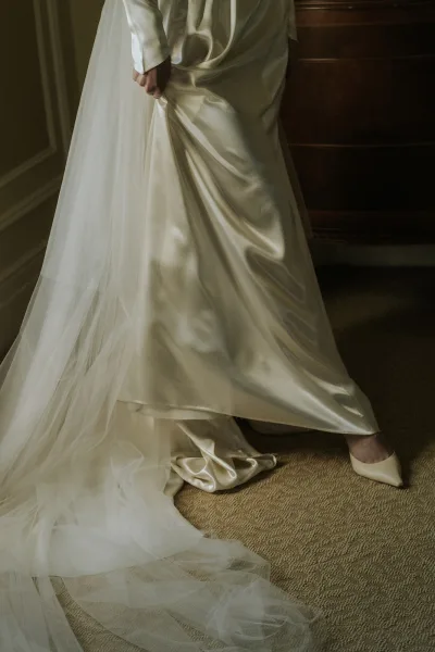Wedding dress close-up of a satin wedding dress, a delicate bridal veil pooling near a wooden dresser on the carpeted floor.