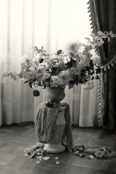 Floral arrangement of urn wedding flowers overflowing from an urn vase before sheer curtains and a tassel curtain on a wood floor.