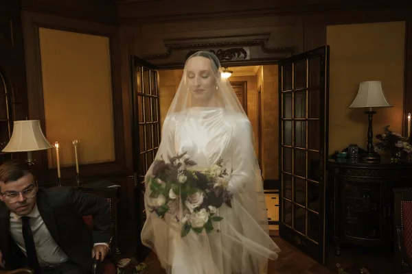 Bridal portrait of a bride with veil in a wood-paneled room, framed by French doors and vintage furniture, serene and softly lit.