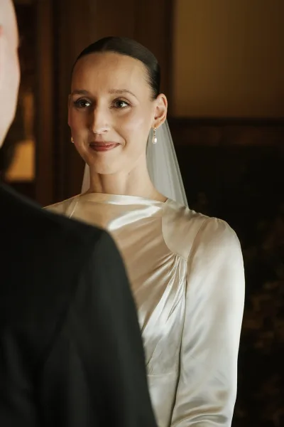 Ceremony moment as a bride in long sleeve gown faces her groom, pearl earrings catching light, in an indoor room with warm wood paneling.