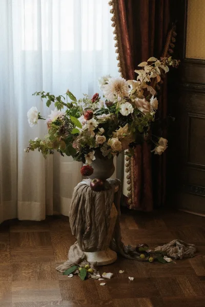 Wedding floral arrangement featuring urn wedding flowers styled with apples, set before red velvet drapes and wood-paneled walls.