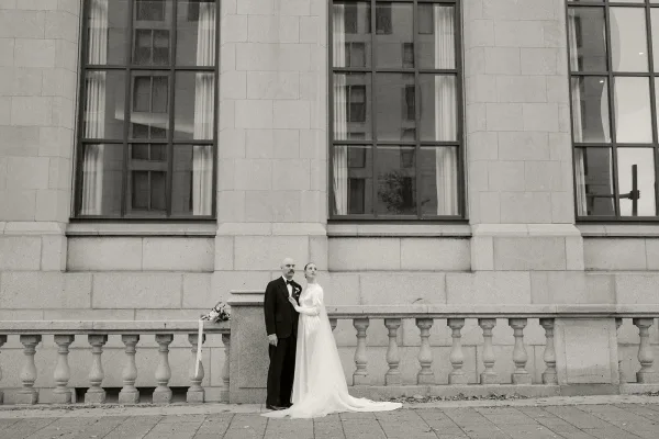 Couple portrait in a black and white wedding photo, bride in a long-sleeve high-neck gown beside the groom, by a stone facade and tall windows.