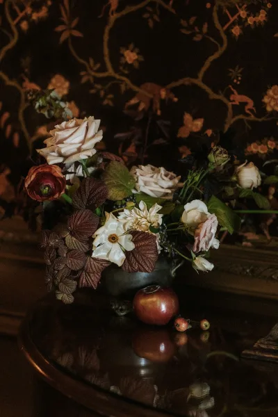 Floral centerpiece, a compote bowl centerpiece with apples, set on a wood table before patterned wallpaper.