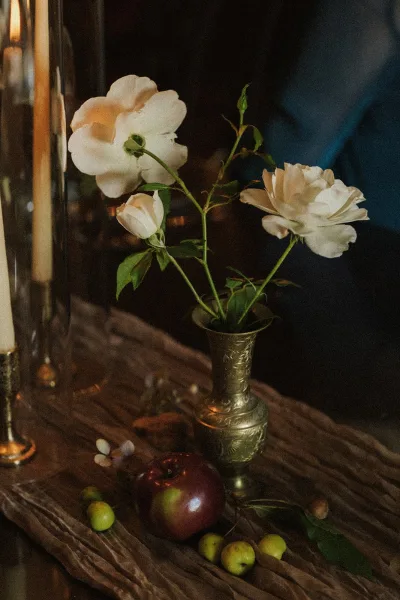 Wedding centerpiece in a brass bud vase with white roses, resting on a wood tabletop within a dark interior, styled for an intimate reception.