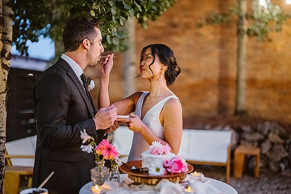 Wedding cake cutting as bride and groom feed each other bites over a white tablecloth with pink flowers, bud vases, candles, and trees behind