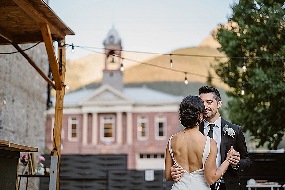 First dance outdoors as bride in a backless wedding dress and groom in black suit sway under string lights in a courtyard at dusk