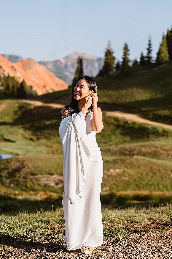 Bridal portrait of a bride adjusting drop earrings in a white cape-sleeve gown on a dirt path with mountains and evergreens behind