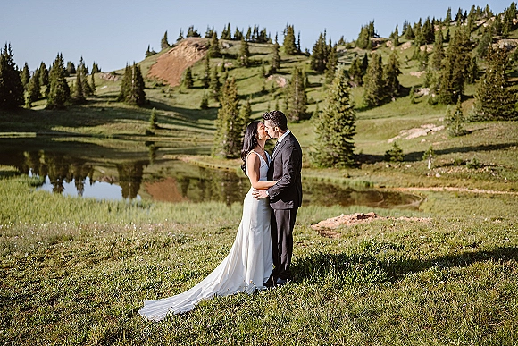 Wedding kiss portrait of bride and groom kissing in a mountain meadow by a lake, her dress train flowing near pine trees