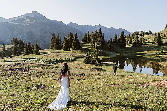 Wedding first look as bride in a satin gown train walks up behind groom in a black suit, with mountains, meadow, and pond beyond