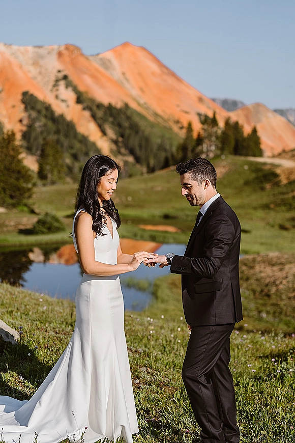 Wedding vows during ring exchange as bride places a ring on groom in a mountain meadow with wildflowers, pond, and pines under blue sky