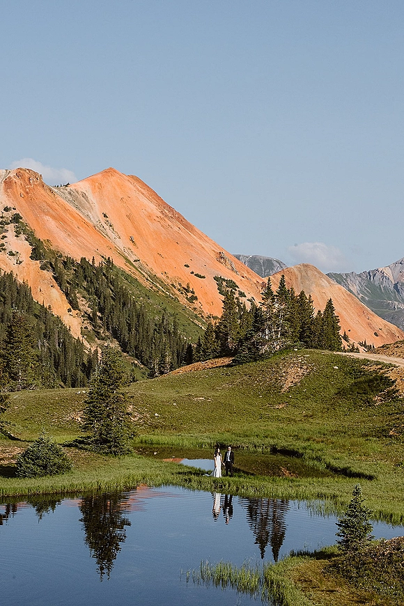 Couple portrait of bride in a wedding dress and groom in a suit standing by a reflective pond in an alpine meadow with pine trees and mountain ridge
