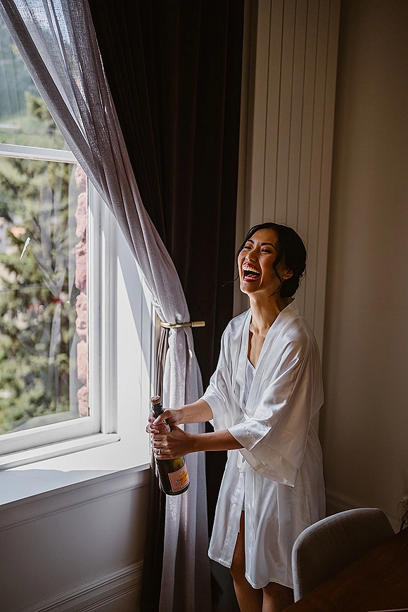 Bride getting ready, bride popping champagne in a white satin robe by a large hotel window, laughing as the cork pops in soft light