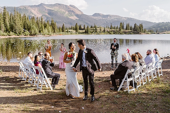 Wedding recessional as newlyweds walk hand in hand down a dirt aisle, bride with bouquet and confetti, lakeside mountains behind guests