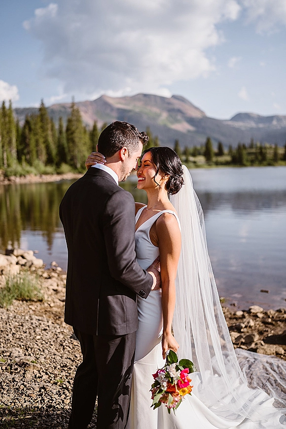 Couple portrait of bride and groom in a mountain wedding portrait, embracing by a lake with evergreens and rocky shoreline under cloudy sky