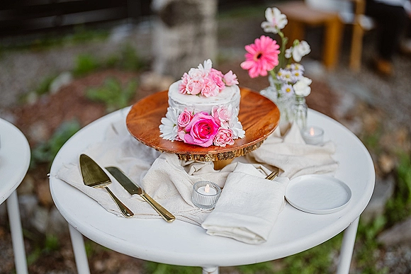 Wedding cake on a rustic wedding cake stand, single tier with pink roses and daisies, styled with candles and cake knife in a garden setting