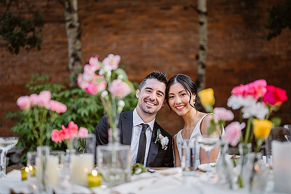 Couple portrait of bride and groom smiling at a candlelit reception table with pink floral centerpieces, garden greenery, and brick wall backdrop