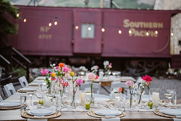 Reception tablescape with wildflower bud vases and roses, taper and pillar candles, gold flatware on a long table by a vintage train car under string lights