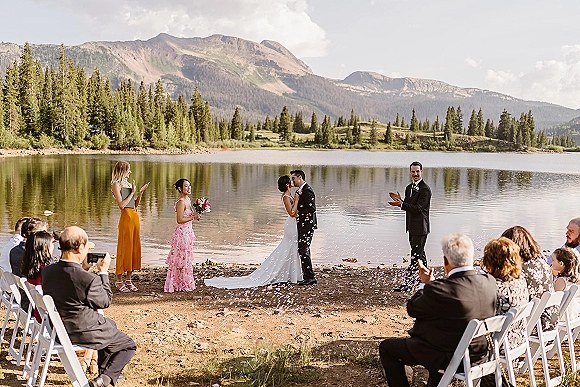 Ceremony kiss at an outdoor wedding ceremony as confetti falls, with lake and mountains behind, bride holding bouquet beside groom in suit