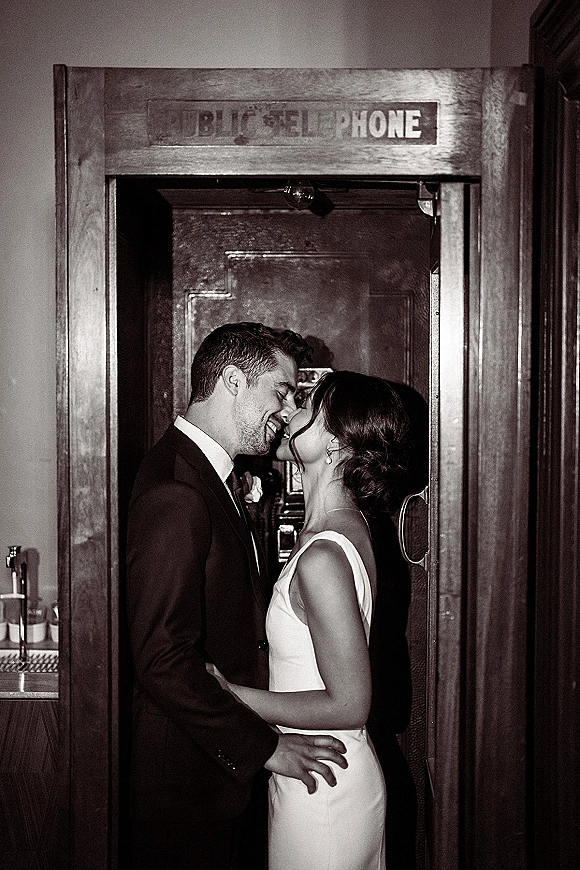 Wedding kiss portrait of newlyweds kissing in profile, bride in simple dress and groom in tuxedo by a vintage telephone booth, wood-paneled wall backdrop