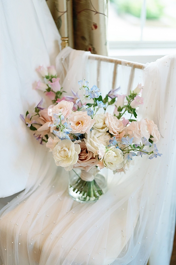 Bridal bouquet of pastel roses and blush flowers with blue delphinium in a glass vase on beaded tulle dress by window light