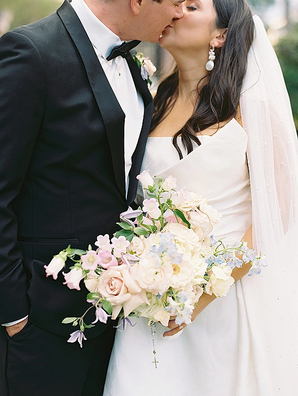 Wedding kiss portrait of bride and groom kissing close up, her veil and blush bouquet visible against soft outdoor greenery backdrop