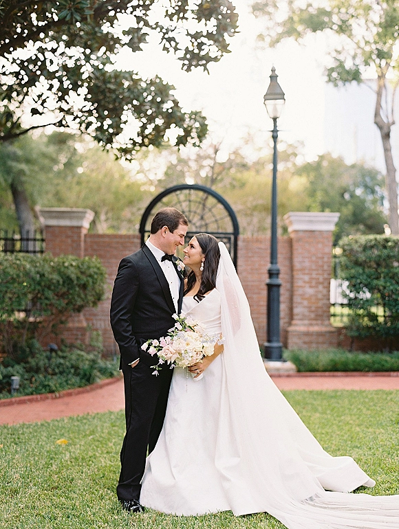 Couple portrait of bride and groom holding hands, bride with bouquet and veil beside a brick wall and iron gate on a garden lawn