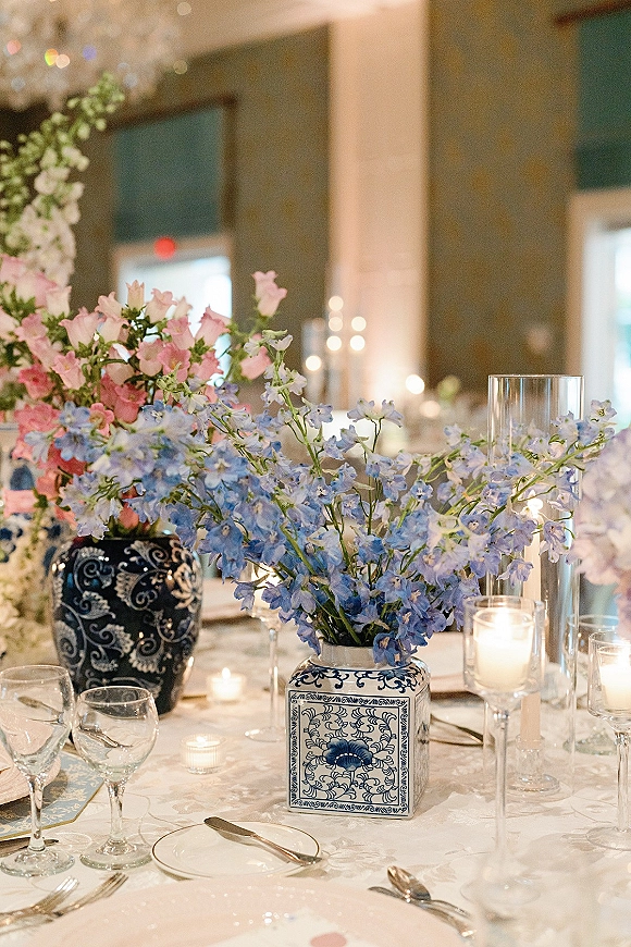 Reception tablescape with wedding table centerpiece in blue and white porcelain vases, taper and votive candles on white linen in a columned hall