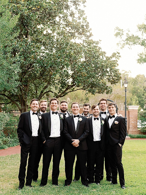 Groomsmen portrait of the groom with groomsmen in black tuxedos and bow ties, standing on a garden lawn by a brick wall and iron fence