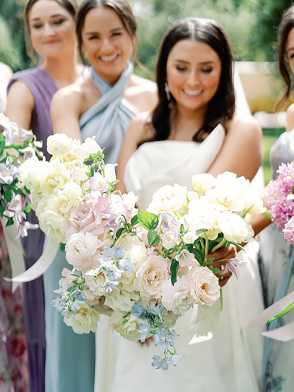 Bridesmaid bouquets of pastel bridesmaid bouquets with blush and ivory roses and blue delphinium, held by the bridal party in sunlit greenery