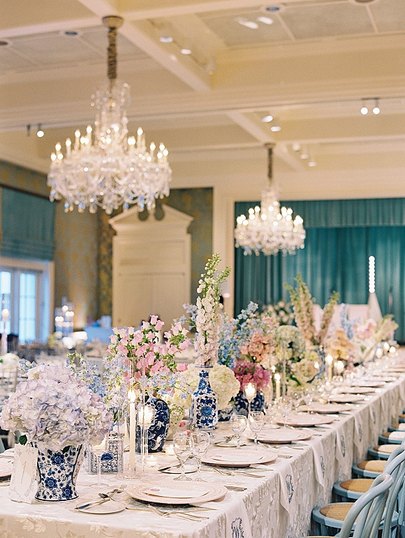 Reception tablescape with wedding head table decor, blue and white porcelain vases of flowers, taper candles, and place settings in a chandelier-lit ballroom