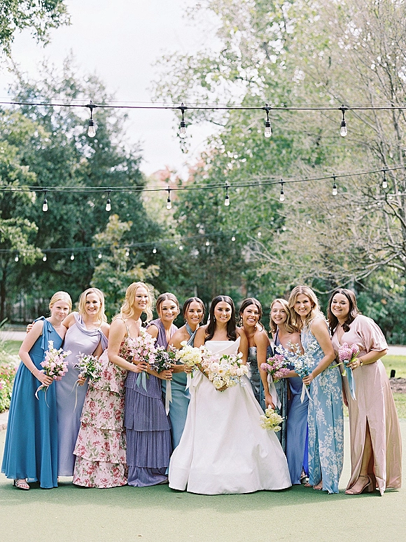 Bridesmaid group photo of the bride with bridesmaids in mismatched pastel dresses holding bouquets with ribbon accents under string lights in a garden lawn