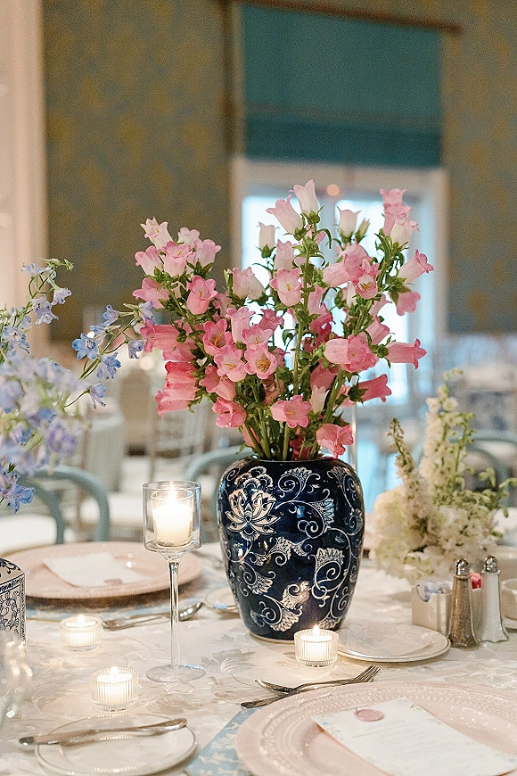 Reception tablescape with wedding table centerpiece in a blue-and-white ceramic vase, pastel flowers, candles, and patterned linens in a dining room