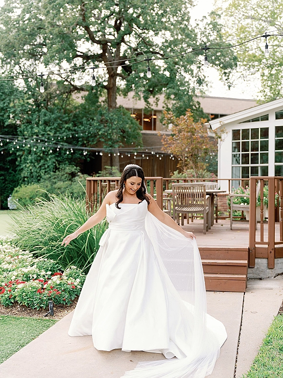 Bridal portrait of a bride in a strapless wedding dress and cathedral veil, holding her train on a garden walkway under string lights