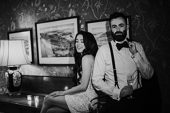 Couple portrait in black and white, bride in strapless sequin dress seated as groom stands with champagne flute in a moody room