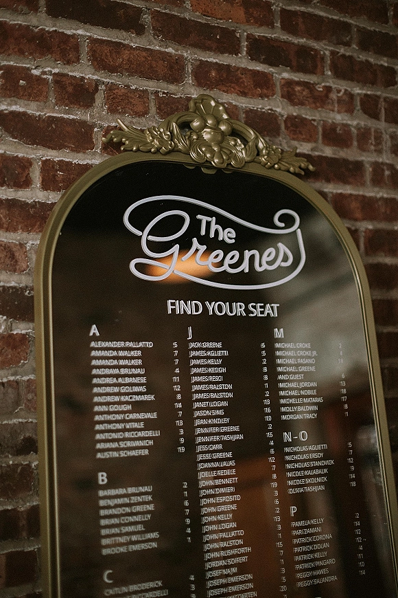 Wedding seating chart on a mirror seating chart sign with white lettering in a gold ornate frame against an exposed brick wall background