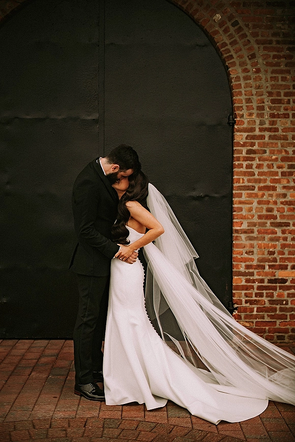Wedding kiss portrait of bride and groom kissing in a close embrace, veil trailing behind, beside a brick wall and arched metal door