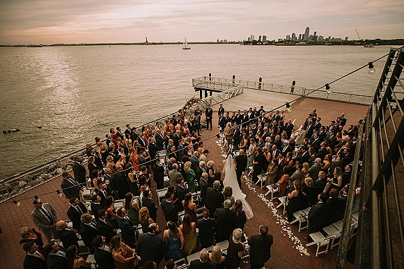 Outdoor wedding ceremony on a waterfront pier with white chairs and petal aisle, bride in cathedral veil under floral arch at sunset