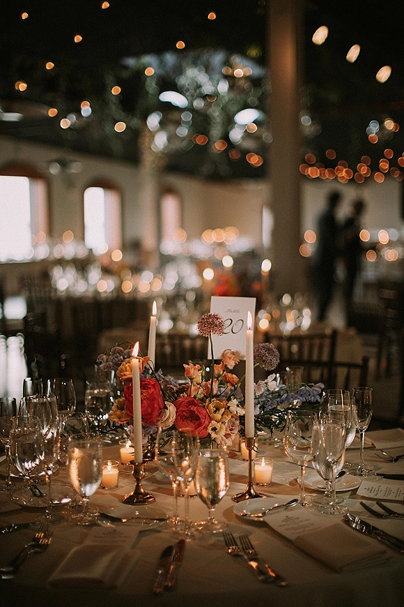 Reception tablescape with a wedding table centerpiece of taper candles in brass candlesticks, florals, and menus under string lights in a beam-lined room