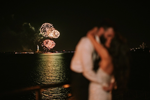 Wedding kiss under fireworks, bride in wedding dress and groom in suit by a waterfront railing with city skyline and statue silhouette at night