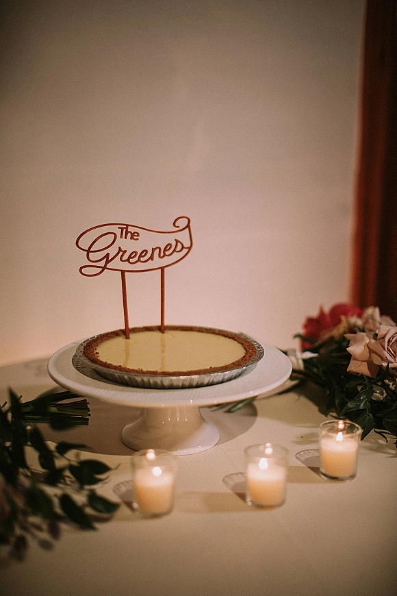 Wedding cake on a wedding dessert table with script topper, pie on a stand, votive candles, greenery garland, and roses on tabletop