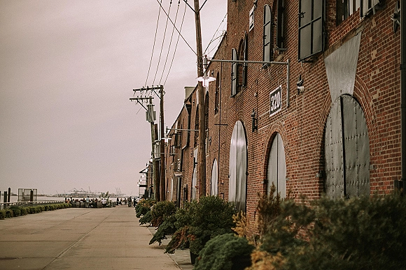 Wedding venue exterior of a brick warehouse building with arched metal doors and wall lamps along a waterfront promenade under overcast sky