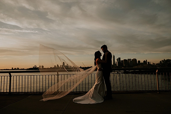 Couple portrait of a bride and groom silhouette embracing by a pier railing, her long veil blowing against a sunset city skyline
