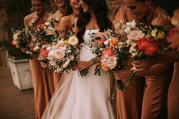 Bridesmaid group photo with bride with bridesmaids laughing, holding rose and greenery bouquets in front of a brick wall outdoors