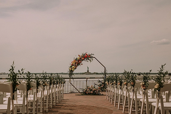 Ceremony setup with an outdoor ceremony aisle leading to a geometric floral arch on a brick patio overlooking the waterfront and sky