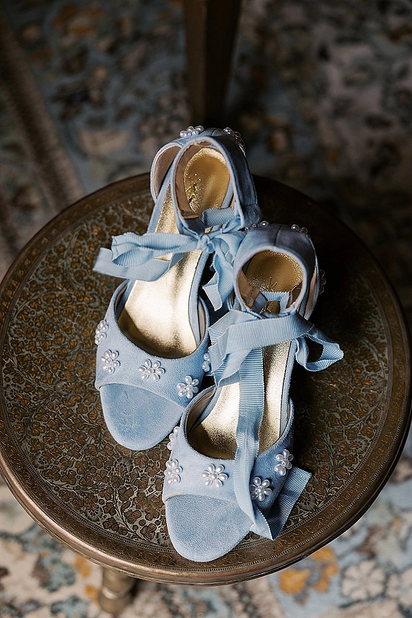 Bridal shoes in baby blue with ribbon ankle ties and pearl embellishments, resting on a round patterned side table over a rug