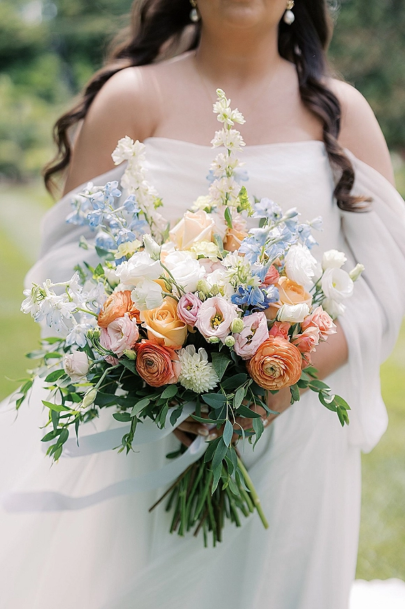 Bridal bouquet of pastel wedding flowers with peach roses, ranunculus, and blue delphinium tied with a white ribbon against greenery lawn background