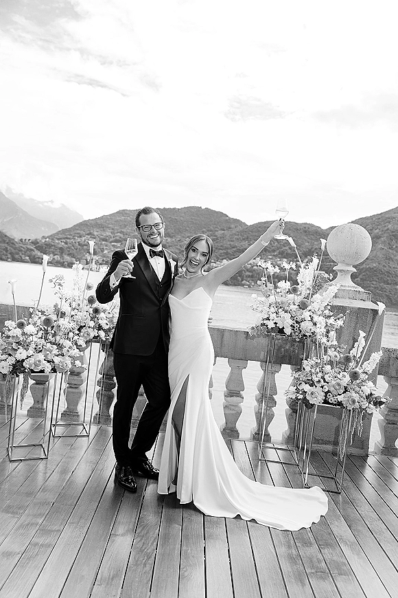 Couple portrait of bride and groom in black tie raising champagne flutes on a stone terrace with lake and mountain views behind