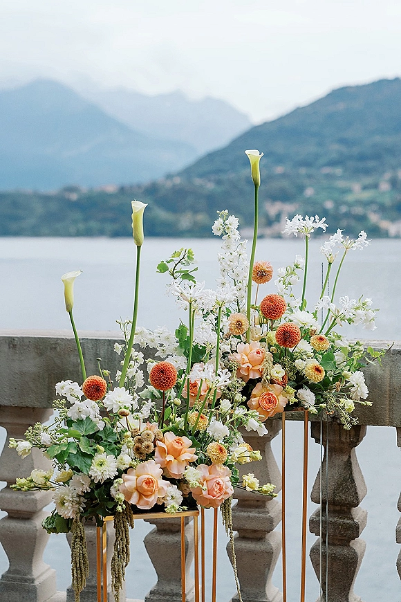 Wedding floral arrangement with ceremony floral installation on gold stands, featuring calla lilies and hanging amaranthus by a stone balustrade lake view