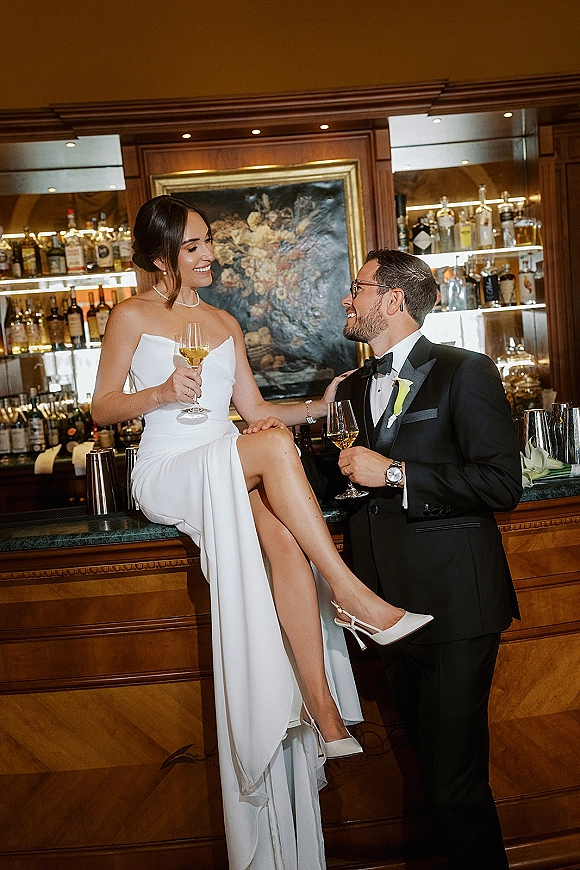 Couple portrait of newlyweds toasting with champagne at a bar, bride in strapless dress on counter beside groom in tuxedo